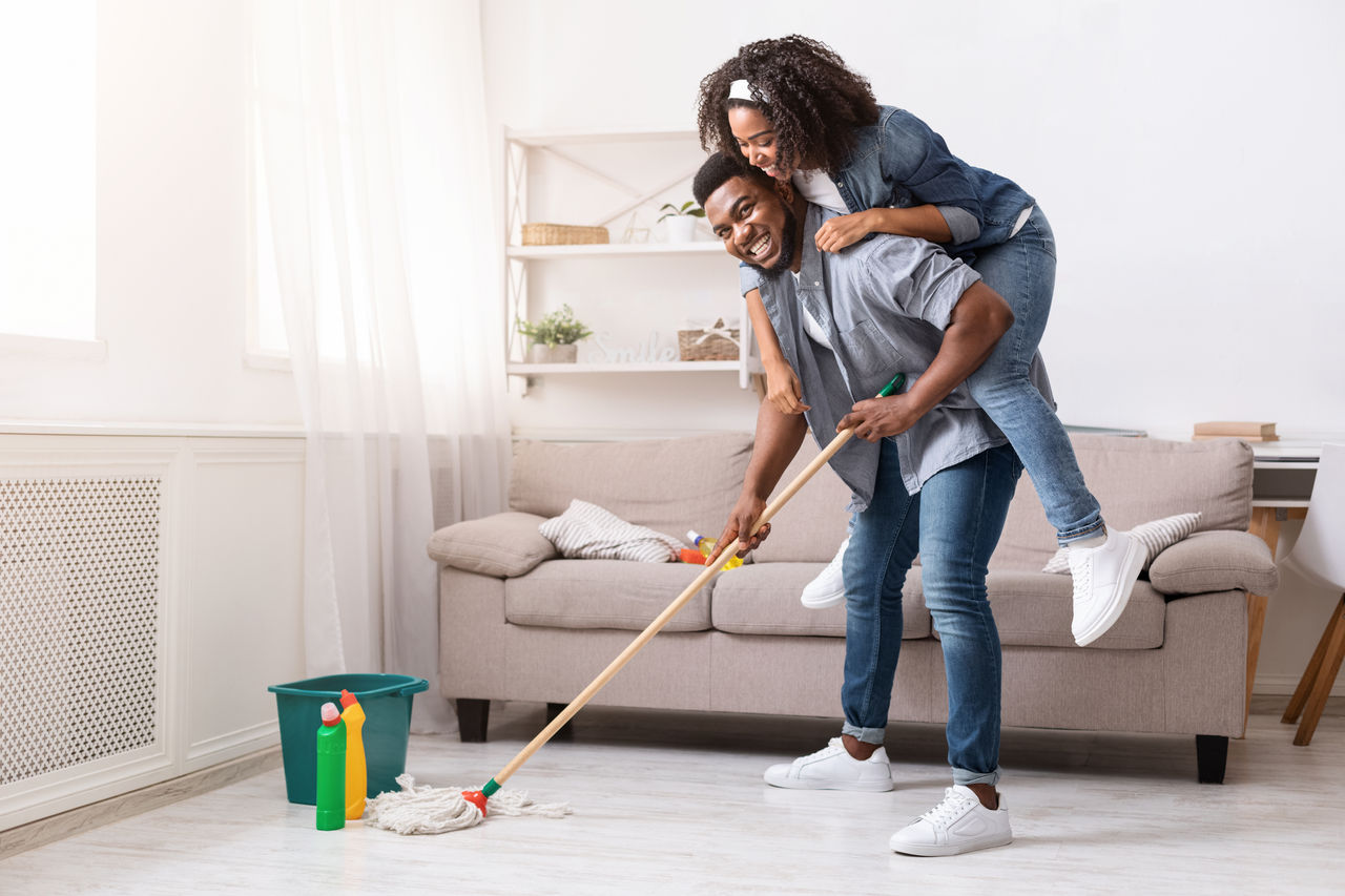 Um homem dá boleia às costas a uma mulher sorridente, enquanto segura uma esfregona numa sala limpa.