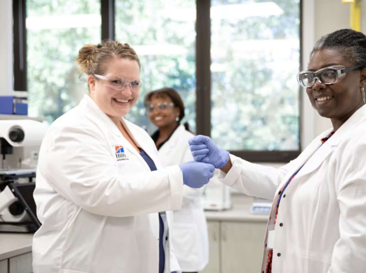 três mulheres sorridentes, cientistas com batas brancas e óculos de proteção