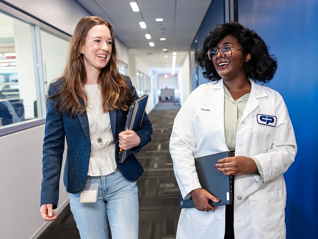 Duas mulheres, uma com roupas casuais e outra com um jaleco branco, caminham e conversam em um corredor de um edifício da Colgate-Palmolive, sorrindo e segurando cadernos.